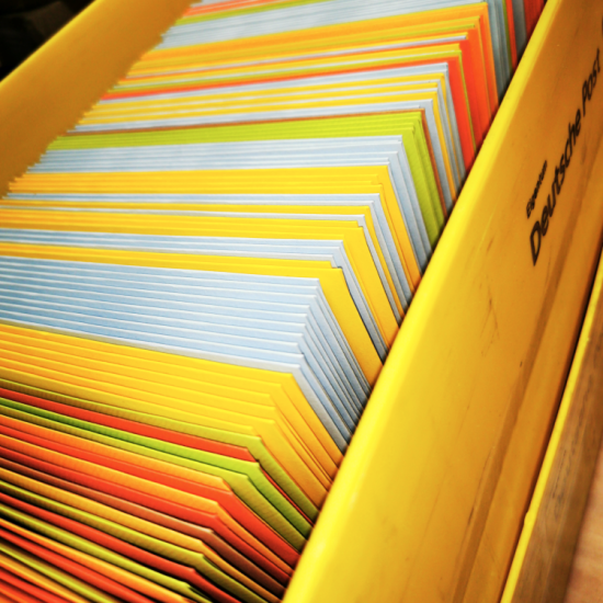 Foto einer gelben Versandbox der Deutschen Post, das vollkommen gefüllt ist mit farbigen gefüllten Briefumschlägen. Foto: Leopoldi-Art